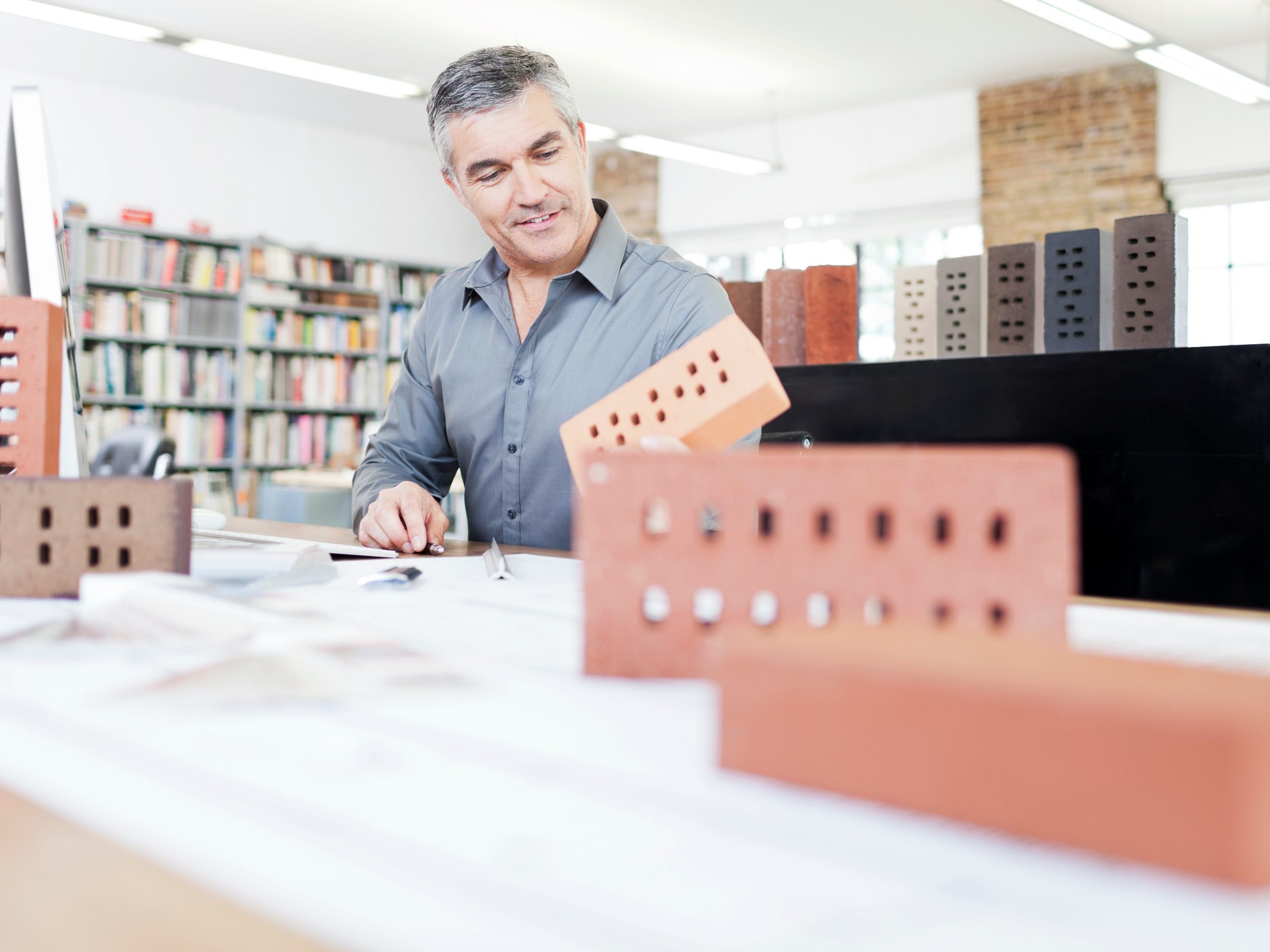 Architekt in seinem Büro sitzt am Schreibtisch mit Blaupause von Ziegeln umgeben