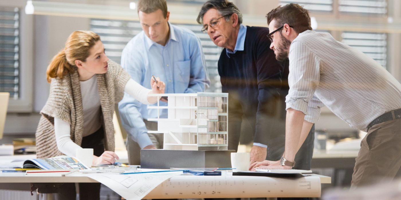 Architect explaining project plan to client in office with the help of an architectural model. Project manager and co-worker standing beside him.