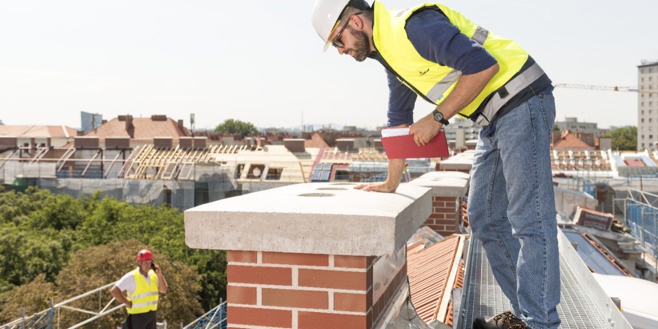 Urban roofers checking brick chimney on roof wearing hard hat and saftey jacket talking on the phone