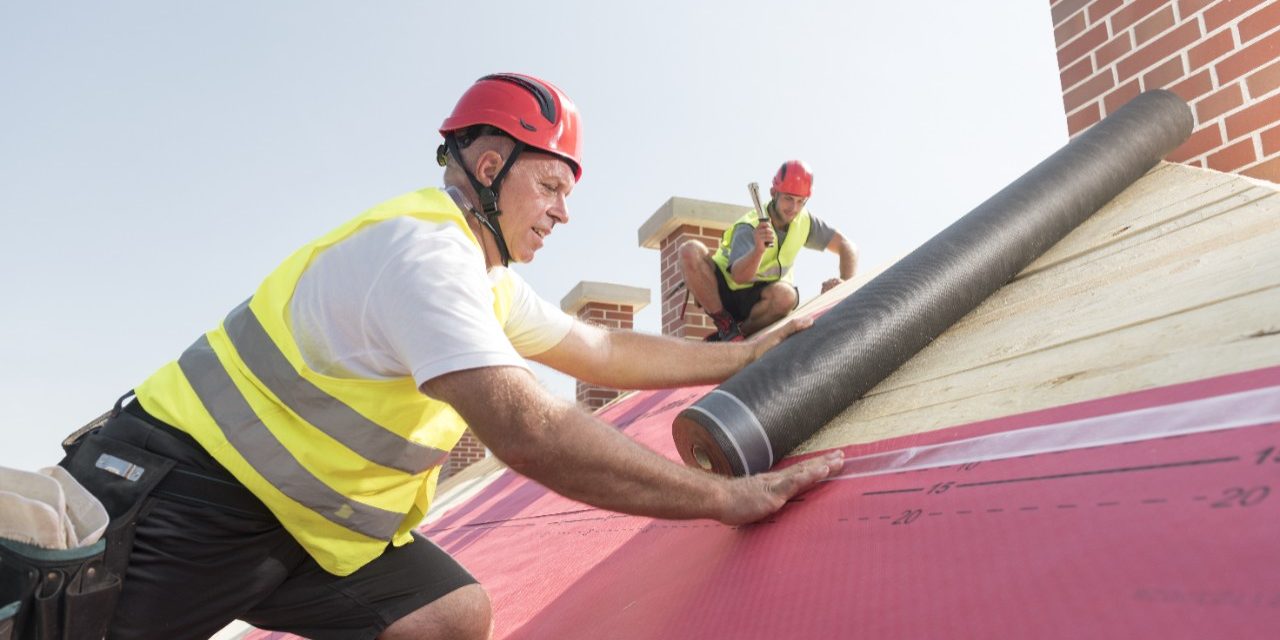 Urban roofers applying roof underlay sheet wearing hard hat and safety jacket brick chimney