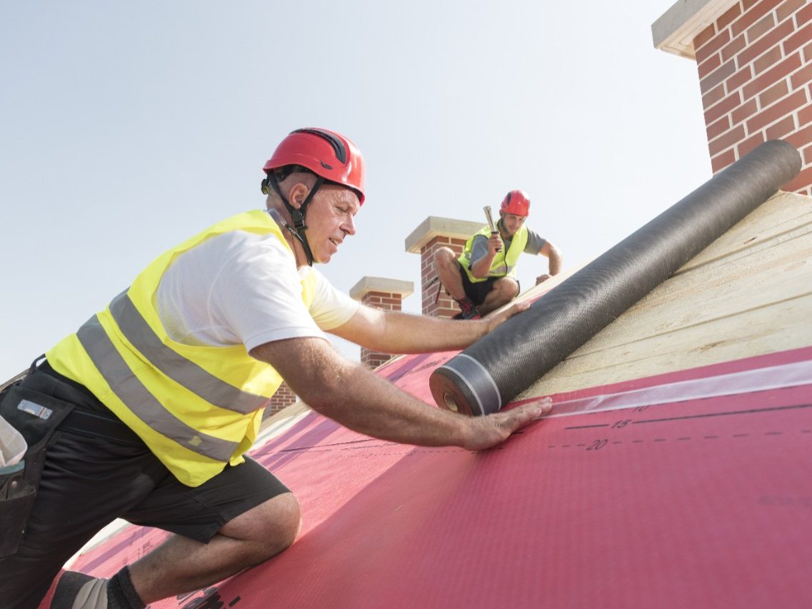 Urban roofers applying roof underlay sheet wearing hard hat and safety jacket brick chimney