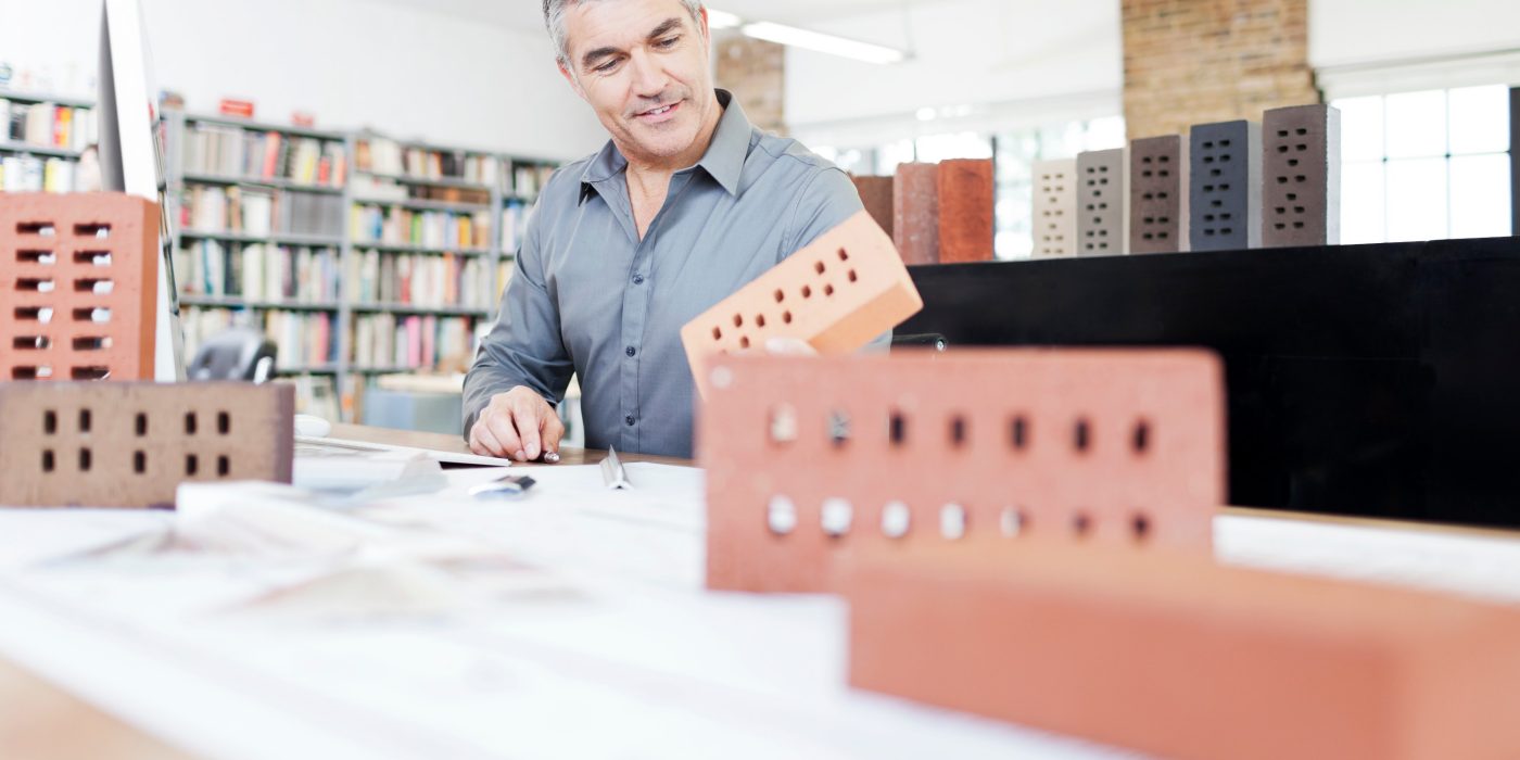 Architekt in seinem Büro sitzt am Schreibtisch mit Blaupause von Ziegeln umgeben