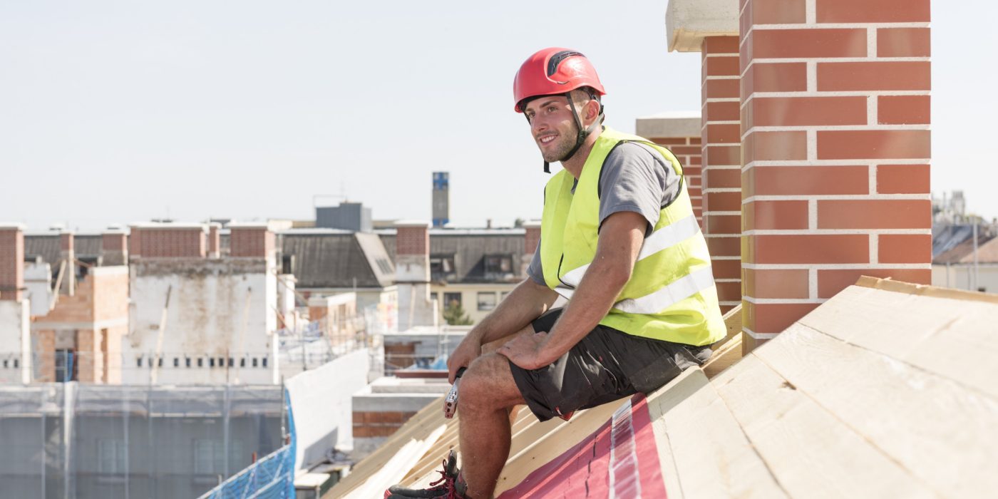 Roofer sitting on roof cover with underlay sheet in front of a chimney in an urban location wearing a hard hat and a safety jacket