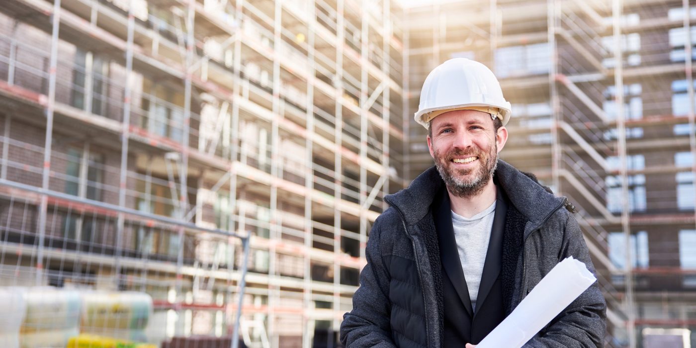 An architect or civil engineer stands with rolled up planning documents in front of a scaffolded apartment building and wears a white hard hat. Photographed in high resolution with copy space