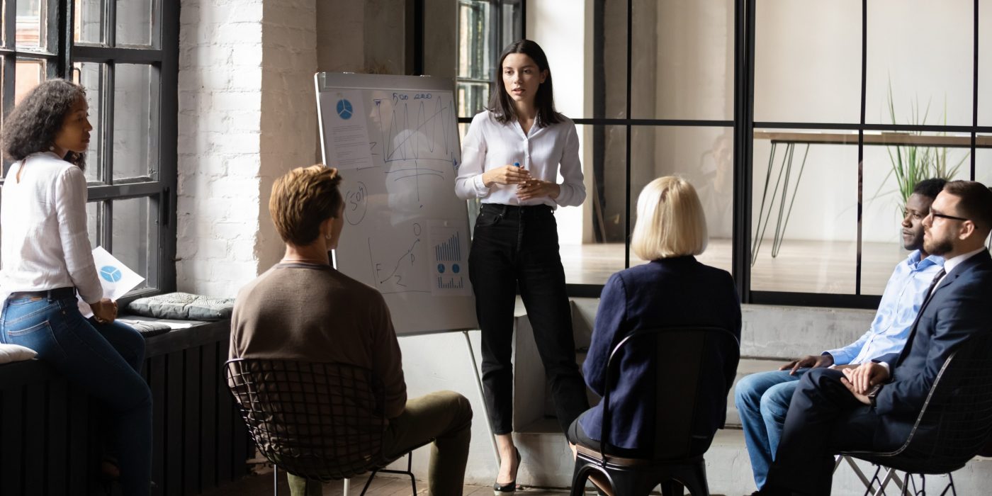 Confident lady business trainer coach leader give flip chart presentation consulting clients teaching employees training team people speaking explaining strategy at marketing workshop concept