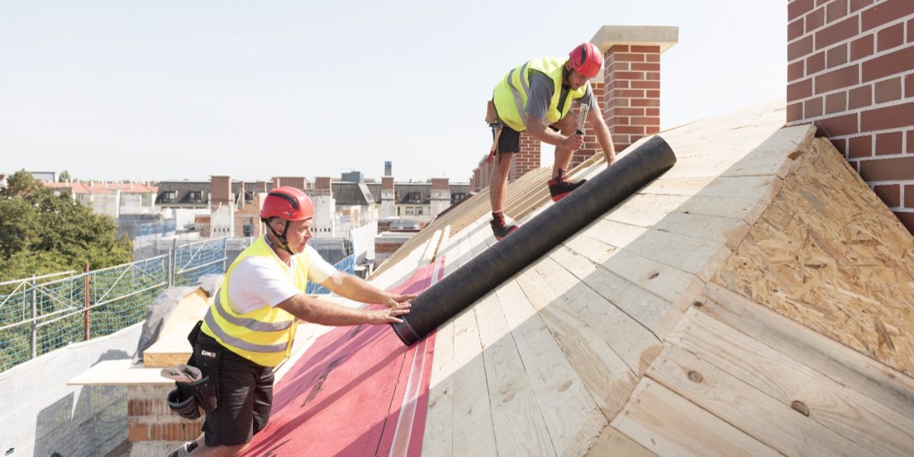 Urban roofers applying roof underlay sheet wearing hard hat and safety jacket brick chimney