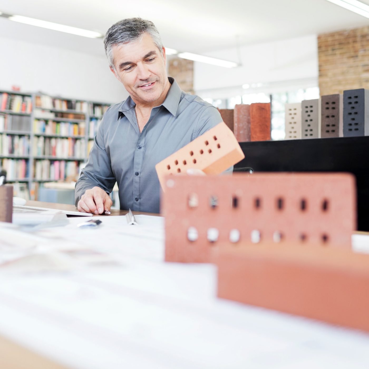 Architekt in seinem Büro sitzt am Schreibtisch mit Blaupause von Ziegeln umgeben