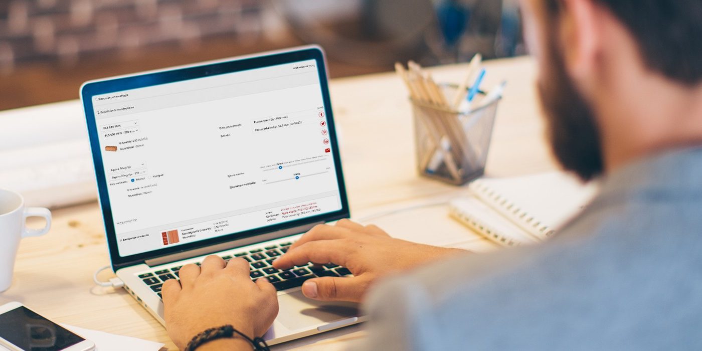 Young man working at modern office space, working on project. Sitting at the table and using laptop. Brick wall in background. Over the shoulder view.