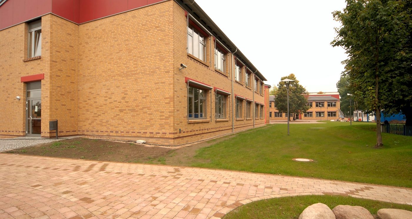 A school with a front garden and trees, you can see the entrance on the left and a sidewalk, Havelland, naturgelb geflammt, glatt, Lüneburg, rotbraun nuanciert, glatt
