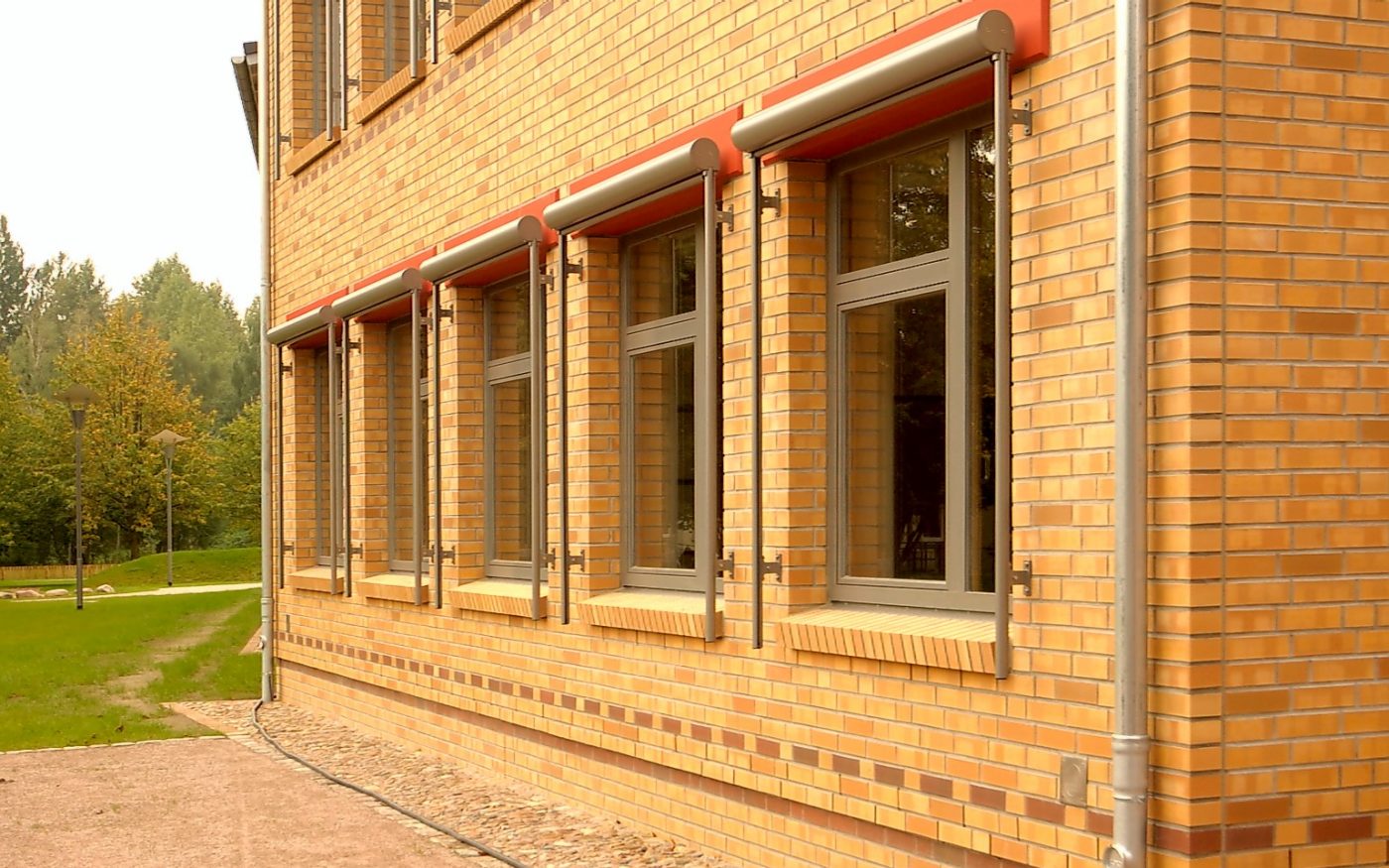 The picture shows a building from an oblique perspective, behind it is a green area with trees. , Havelland, naturgelb geflammt, glatt, Lüneburg, rotbraun nuanciert, glatt