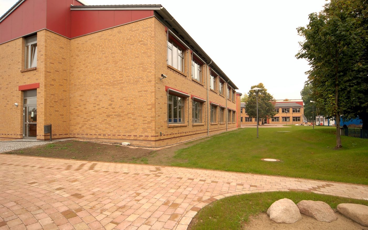 A school with a front garden and trees, you can see the entrance on the left and a sidewalk, Havelland, naturgelb geflammt, glatt, Lüneburg, rotbraun nuanciert, glatt