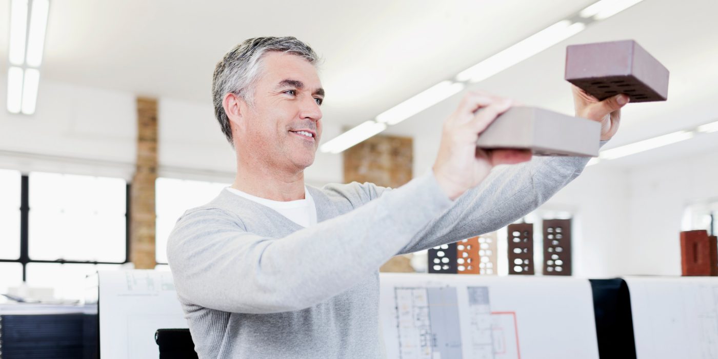 Lächelnder Architekt in seinem Büro, umgeben von Bauplänen, der Ziegelsteine prüft