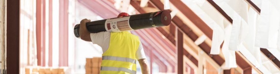 Urban roofer carrying underlay sheet on his shoulder in renovated attic wearing hard hat and safety jacket