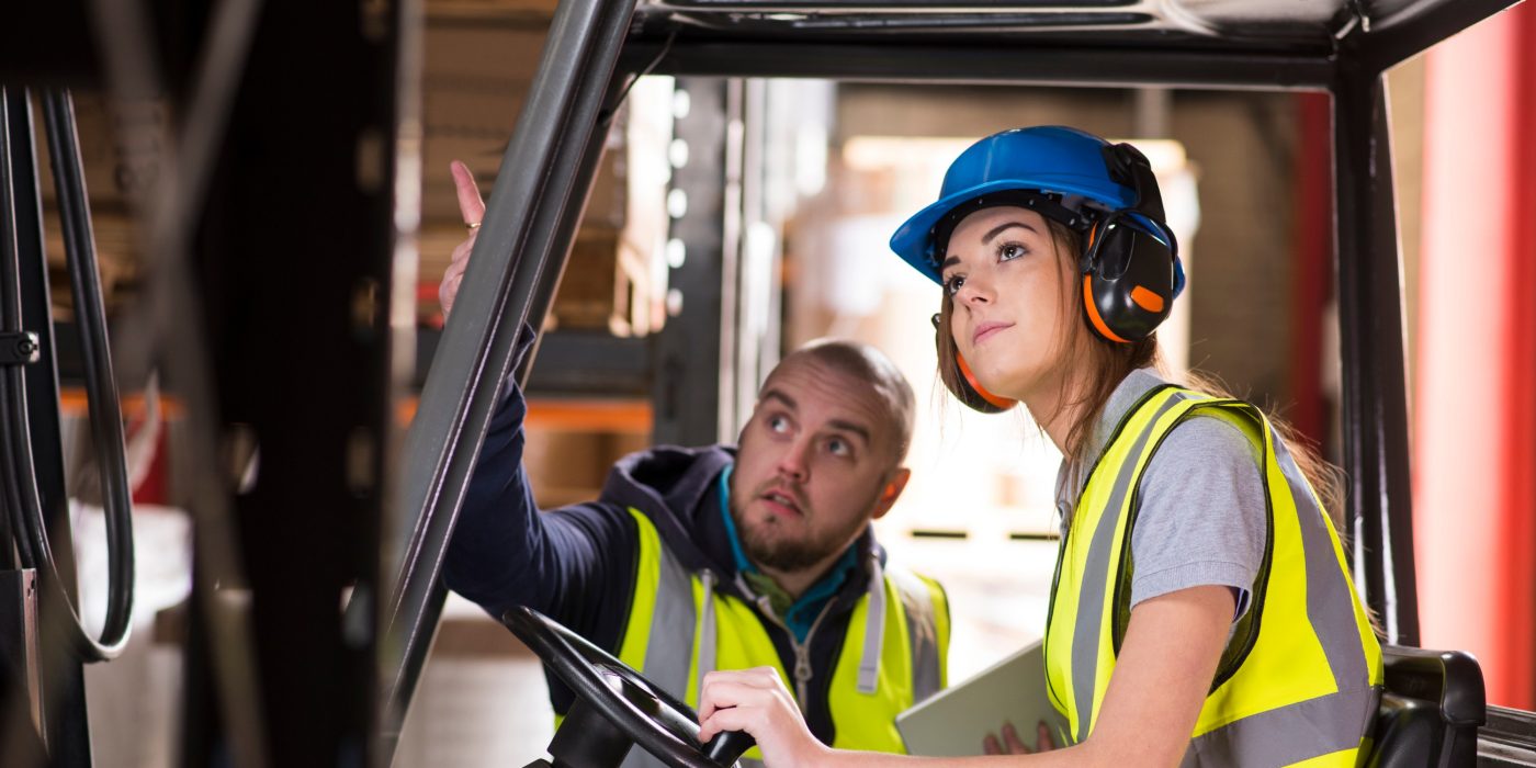 A young female forklift driver is being trained on approaching the racking of the warehouse . A male colleague or trainer can be seen advising her on how to perform the manoeuvre . She is wearing a hi vis jacket , hard hat and ear defenders .