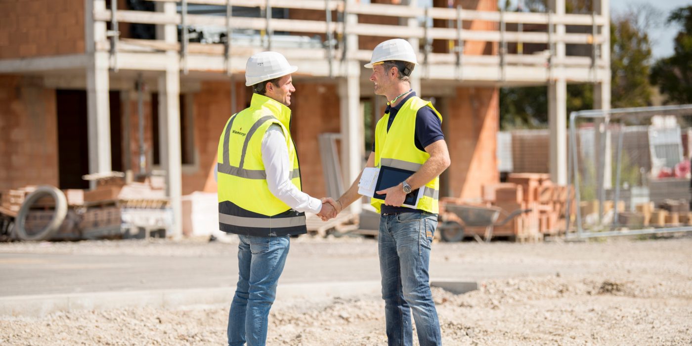 Manager and builder shaking hands on construction site in front of building shell